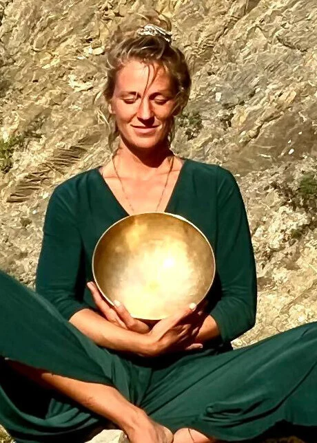 Laura Nolte sitzt im Schneidersitz vor einer Felswand und hält eine goldene Klangschale im Schoß.